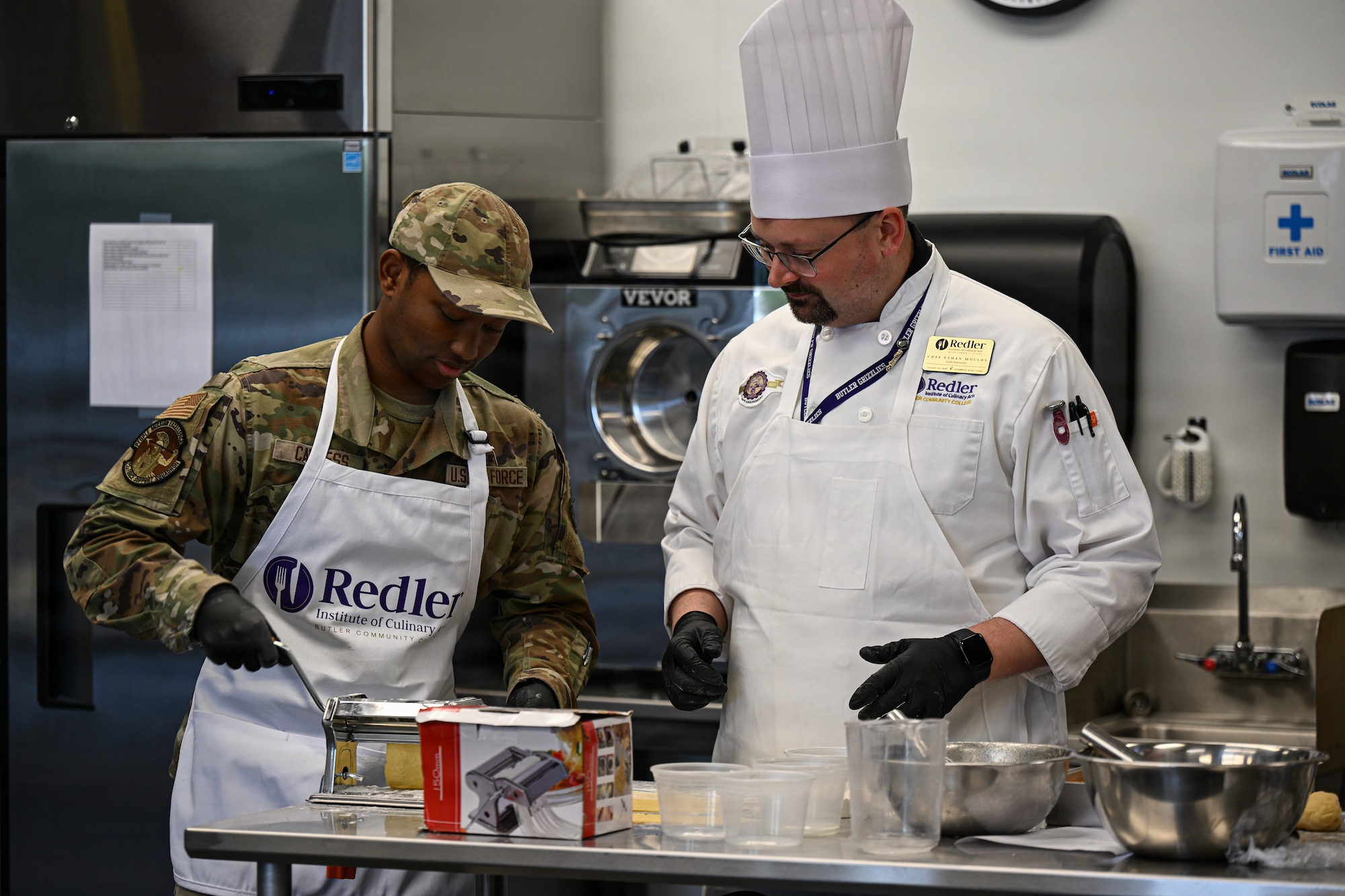 Senior Airman Kamar Cavness, 22nd Force Support Squadron fitness and sports journeyman, and Chef Ethan Moulds, Redler Institute of Culinary Arts head instructor, use a manual appliance to roll out pasta dough at the Redler Institute of Culinary Arts in Andover, Kansas, April 28, 2026. Ethan walked around providing guidance to the Airmen who were learning new cooking techniques during a partnered program between Redler and McConnell  Air Force Base. (U.S. Air Force photo by Senior Airman Amelio Brown)