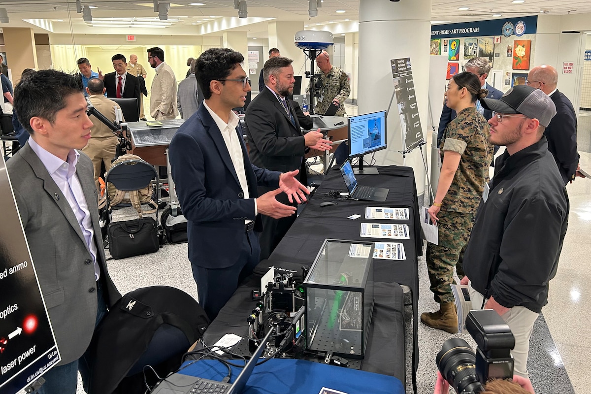 Several men stand on one side of a table talking to people on the other side of the table. On the table are computers and an array of equipment featuring lasers and optical gear.