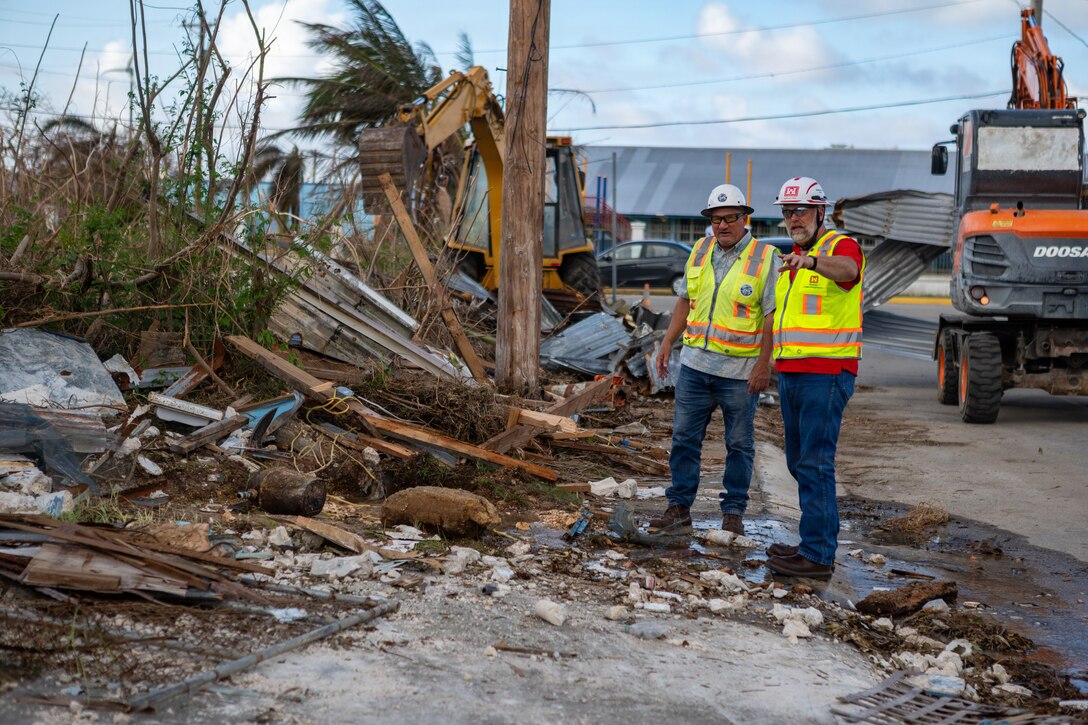 Two men in safety vest stand on the side of the road next to piles of storm debris.