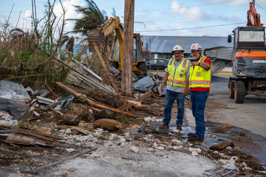 Two men in safety vest stand on the side of the road next to piles of storm debris.