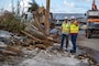 Two men in safety vest stand on the side of the road next to piles of storm debris.