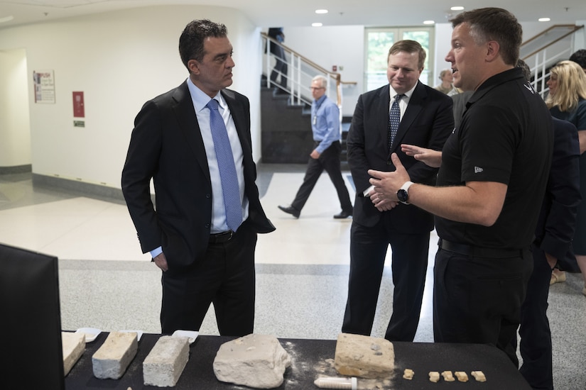 Three men engage in conversation. They stand near a tabletop display of what appears to be rocks and bricks.