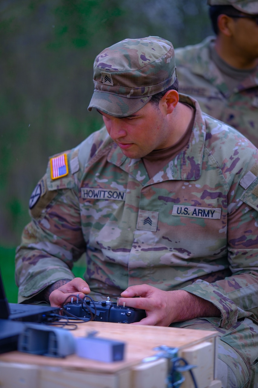 A man wearing camouflage military uniform looks at a laptop as he holds a remote control. Another man in similar attire is in the background.