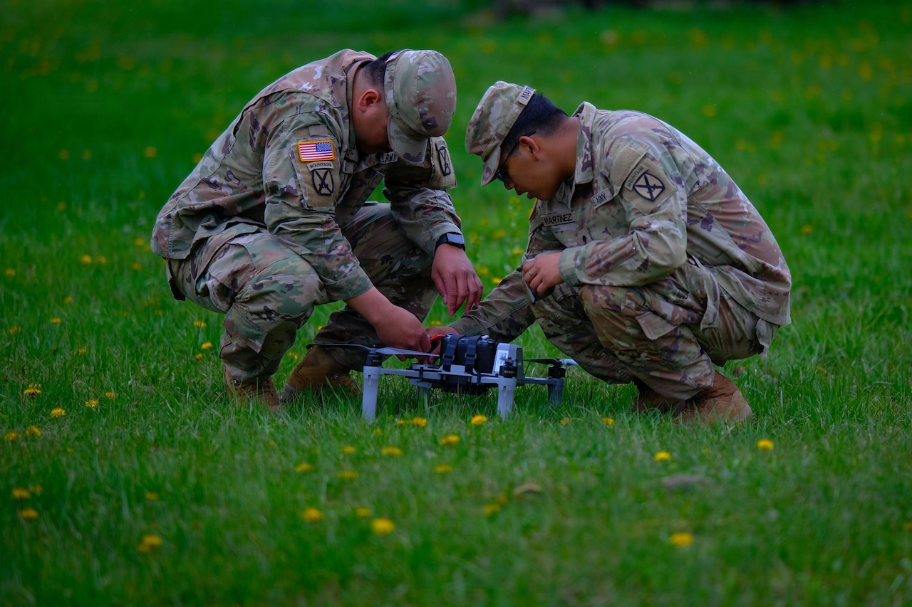 Two men wearing camouflage military uniforms crouch down in a field to work on a military drone.