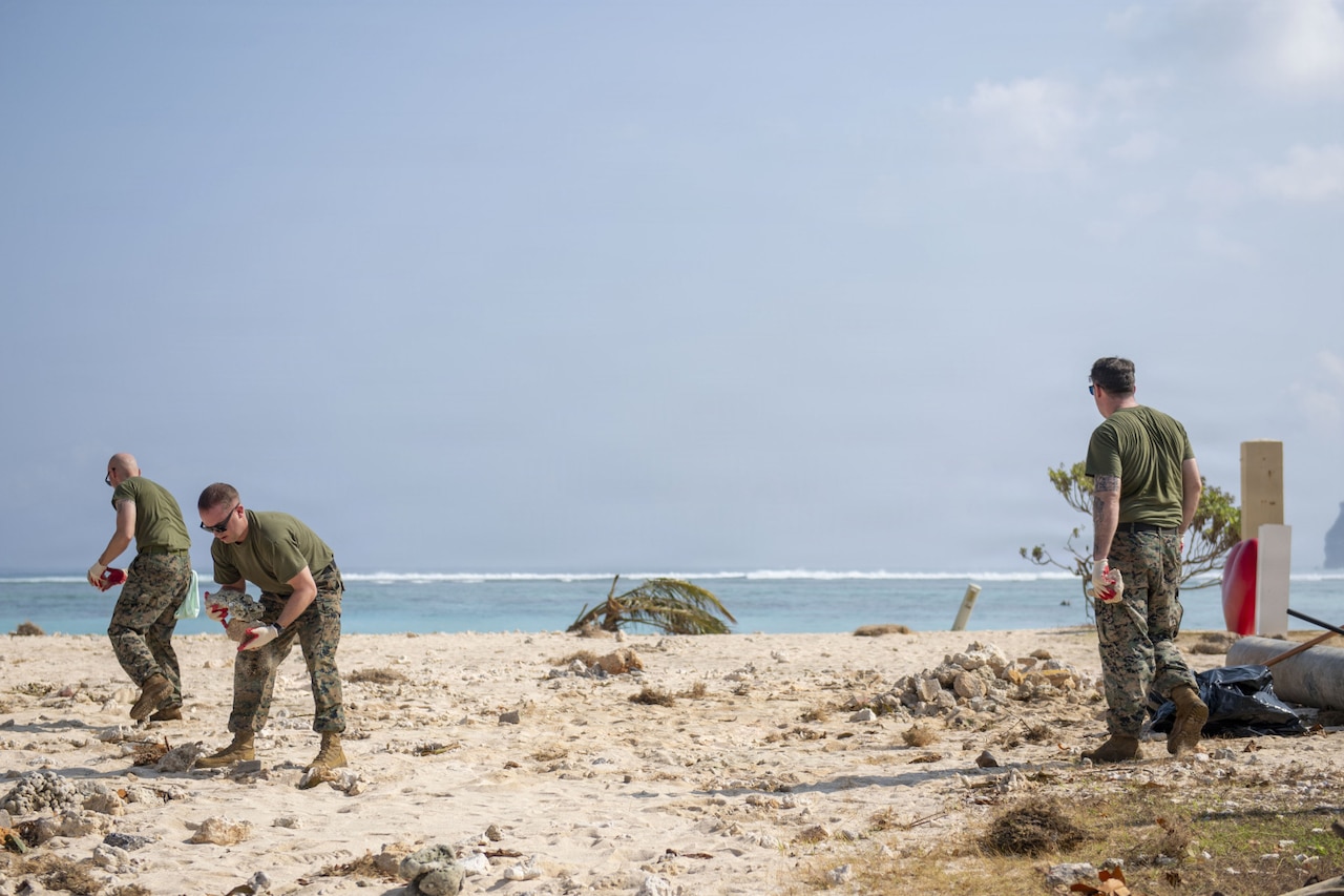Three men in camouflage military uniforms pick up debris from a beach.