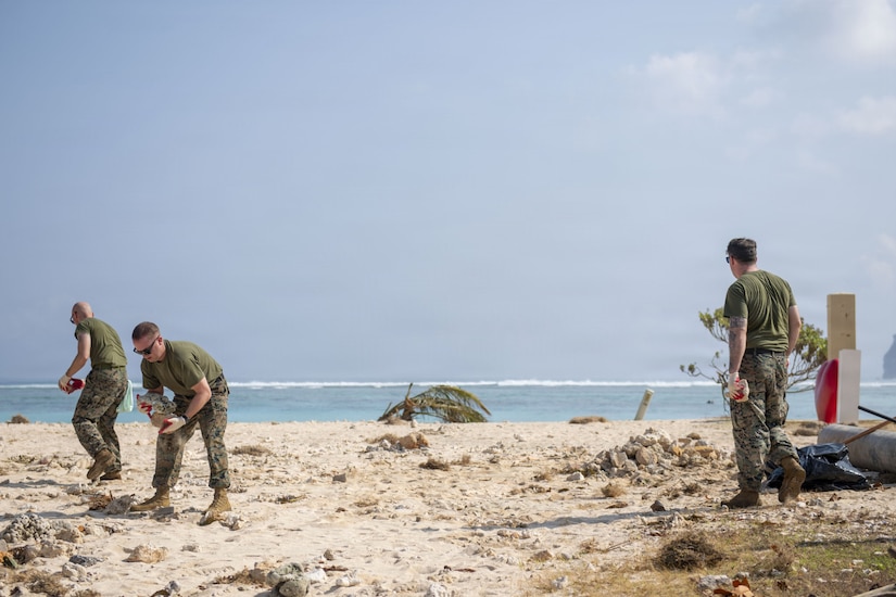 Three men in camouflage military uniforms pick up debris from a beach.