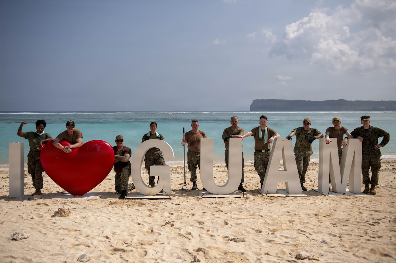 Ten people in camouflage military uniforms pose for a photo on a beach around a sign that reads, "I love Guam."
