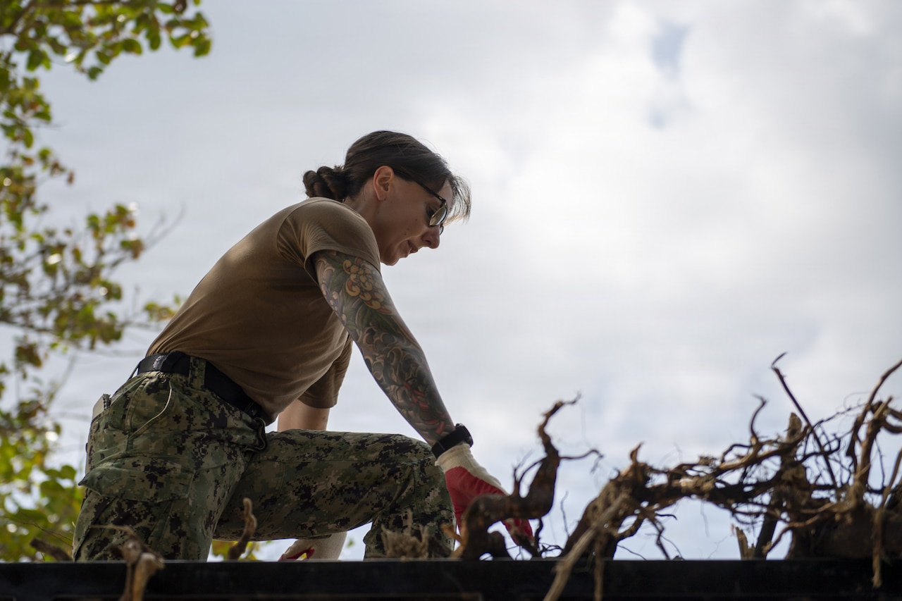 A woman wearing a camouflage military uniform and sunglasses stands on top of debris in the back of a cargo vehicle.