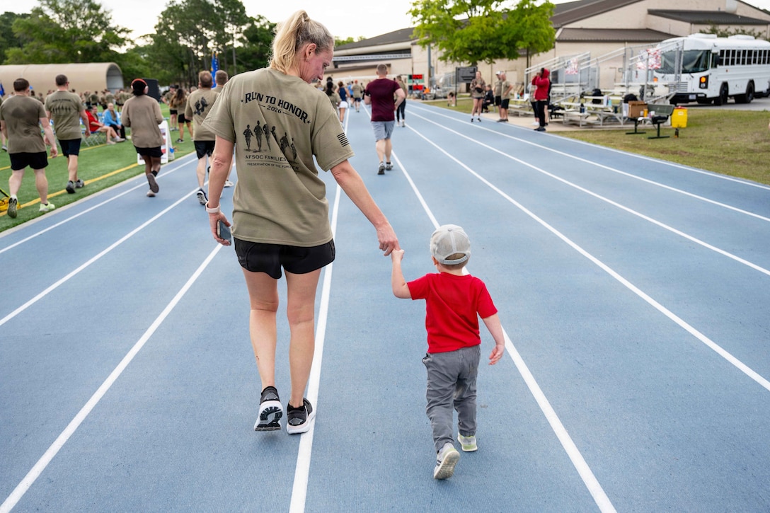 An airman in running gear holds hands with a toddler while walking on a blue track with fellow airmen and family members walking nearby.