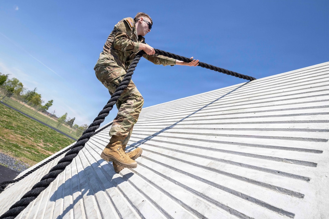 A soldier holds black rope while scaling a white wall next to a field against a blue sky.