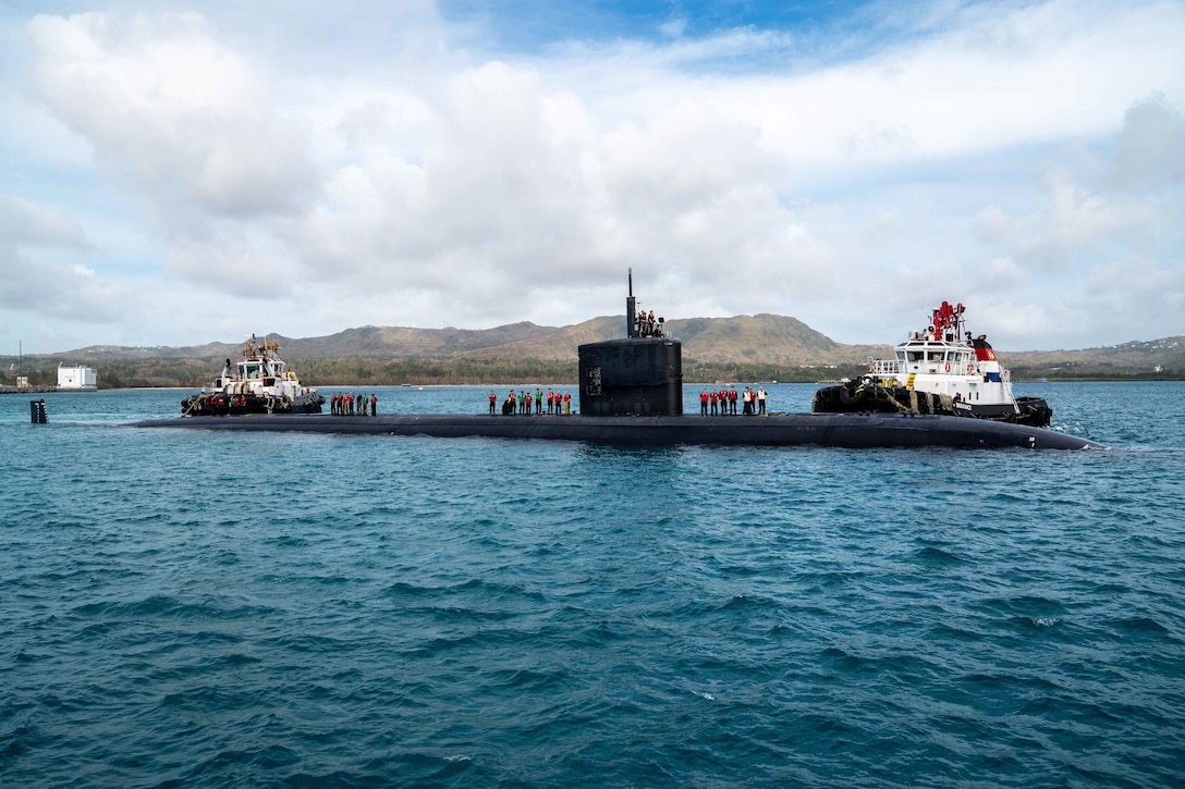 Two boats approach opposite ends of a submarine in blue water as three groups of sailors stand on top of it in front of an island.