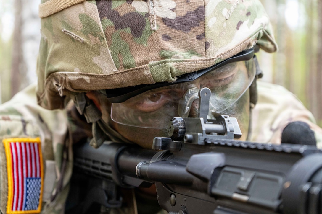 A close-up of a solder in camouflage lying in the woods looking through the scope of a weapon against a blurred background.