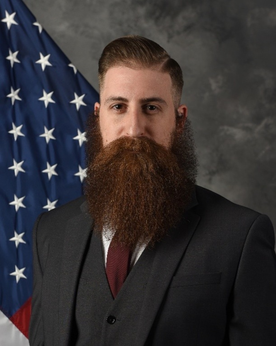 Mr. Cross poses for a photo, pictured from the waist up and the U.S. flag in the background and gray backdrop.