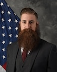 Mr. Cross poses for a photo, pictured from the waist up and the U.S. flag in the background and gray backdrop.