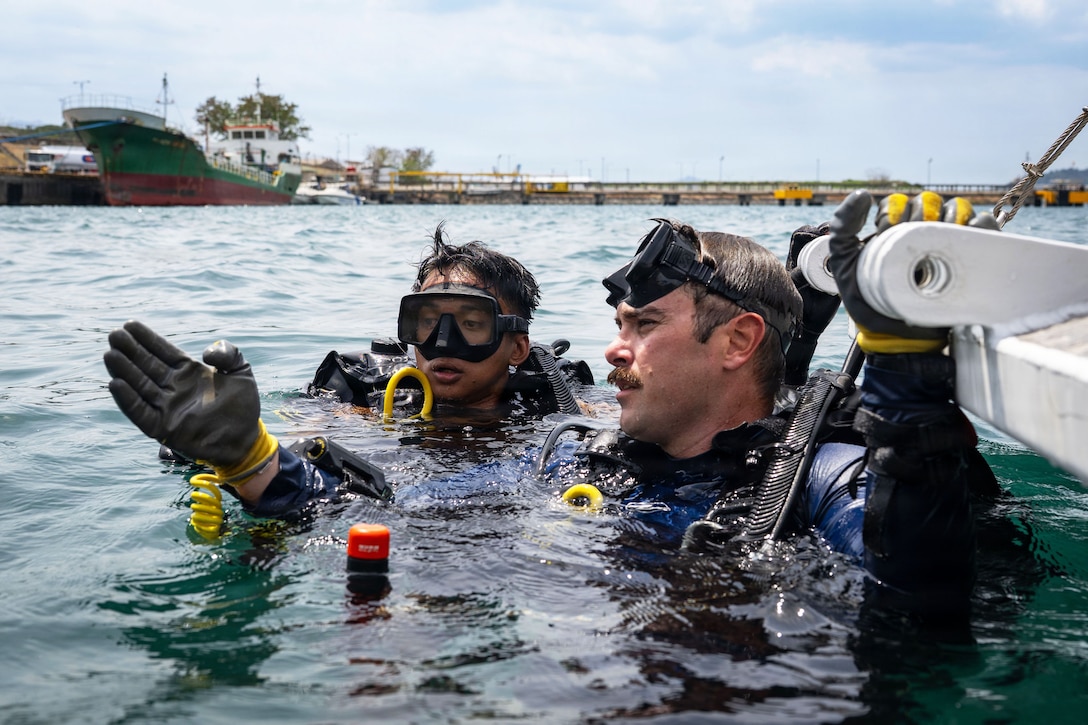 Two divers with goggles on their heads are in a bay with their heads and shoulders above water as one diver holds out a hand as if explaining information.