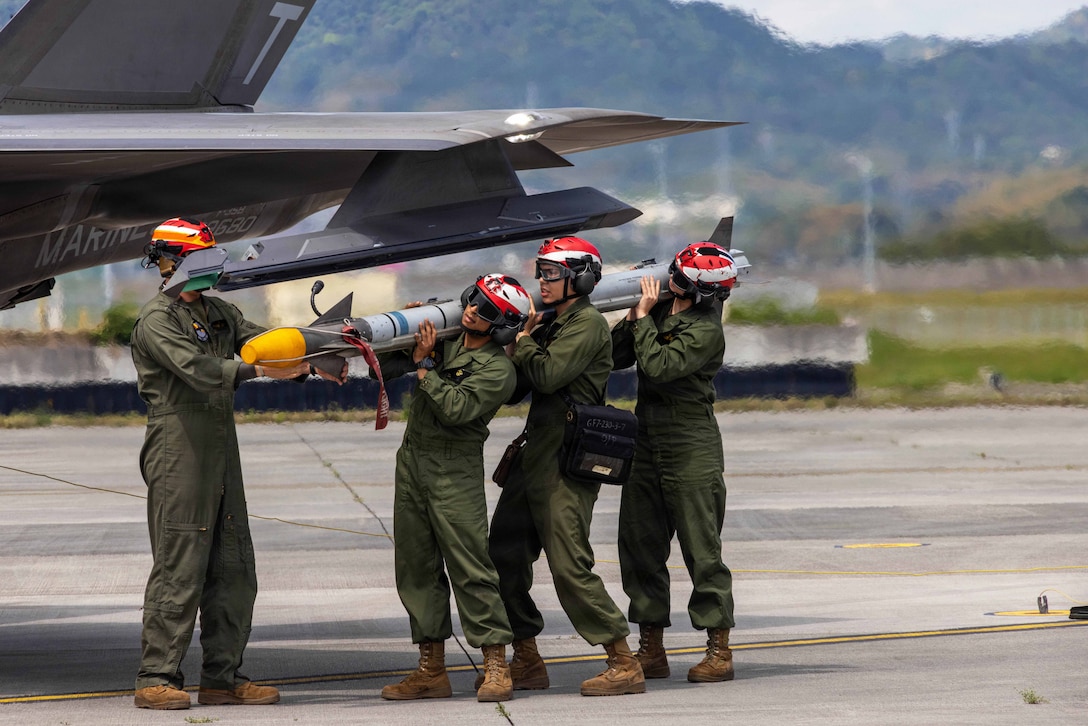 Marines wearing green jumpsuits and red, white and yellow helmets hold up a missile near the wing of a jet on a tarmac, with mountains in the background.