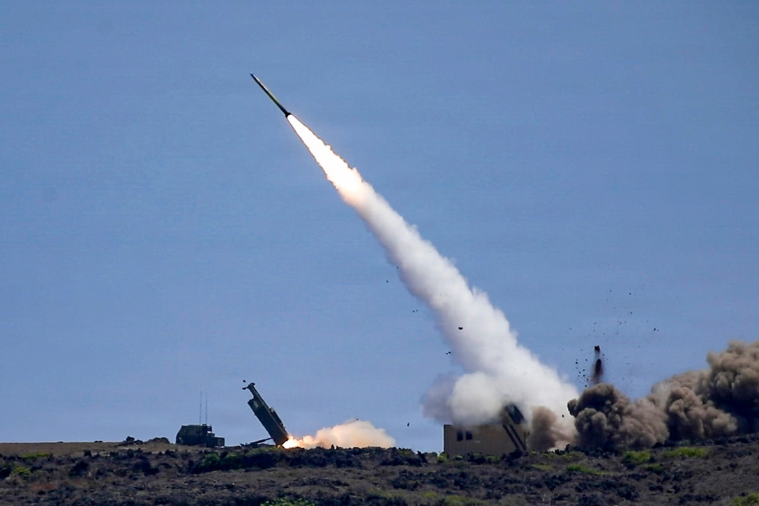 A missile has been fired into the air and is above a grass-covered ground, with fire and smoke behind it.
