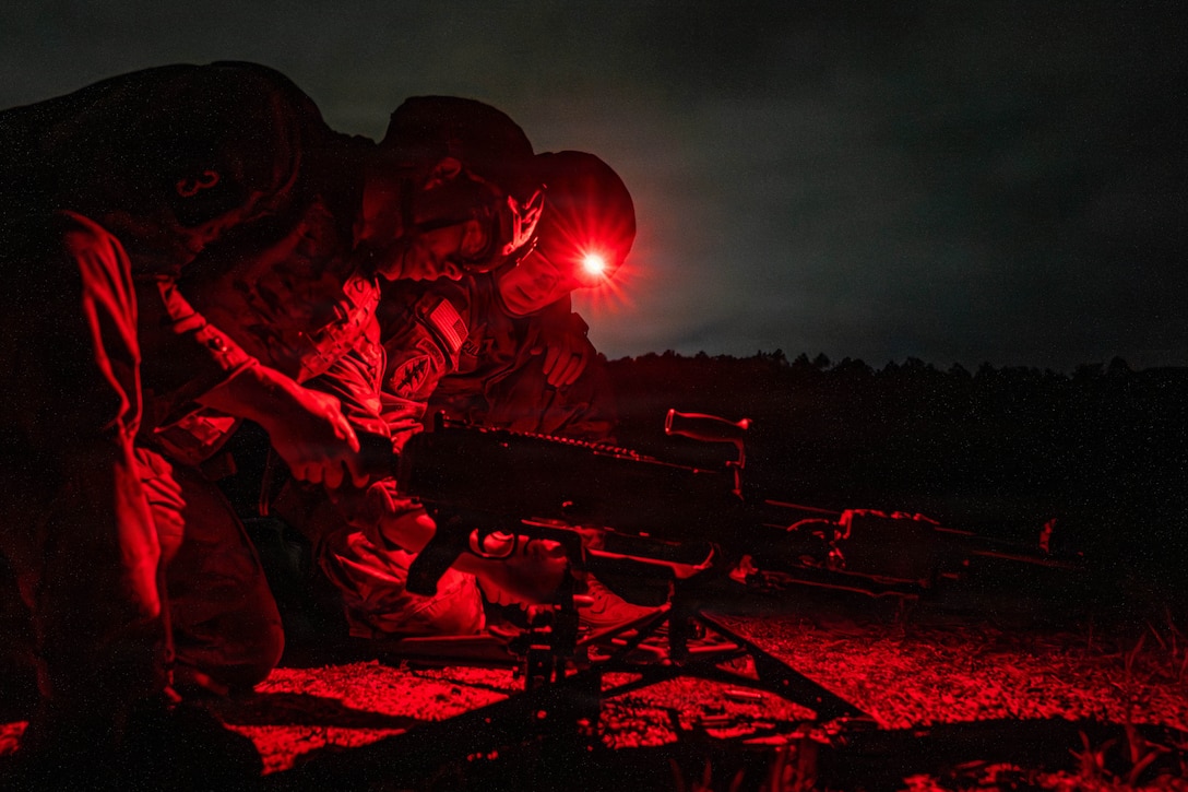 Two soldiers prepare to fire a machine gun at night, with one touching the end of the gun as it rests on a mount on the ground and the other shining a red helmet light on it.