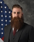 Mr. Cross poses for an official photo with a U.S. flag and grey blackdrop.