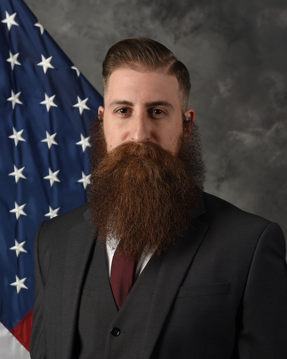 Mr. Cross poses for an official photo with a U.S. flag and grey blackdrop.