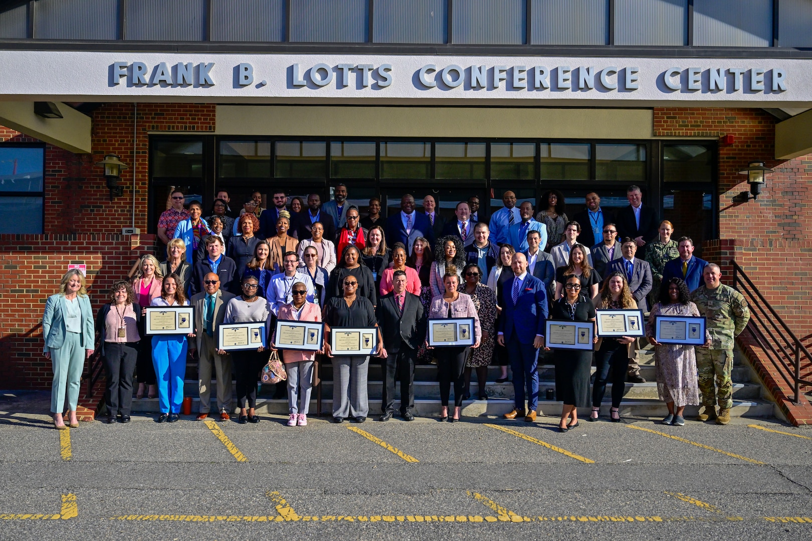 A large group of employees stand on stairs in front of a building holding their certificates.