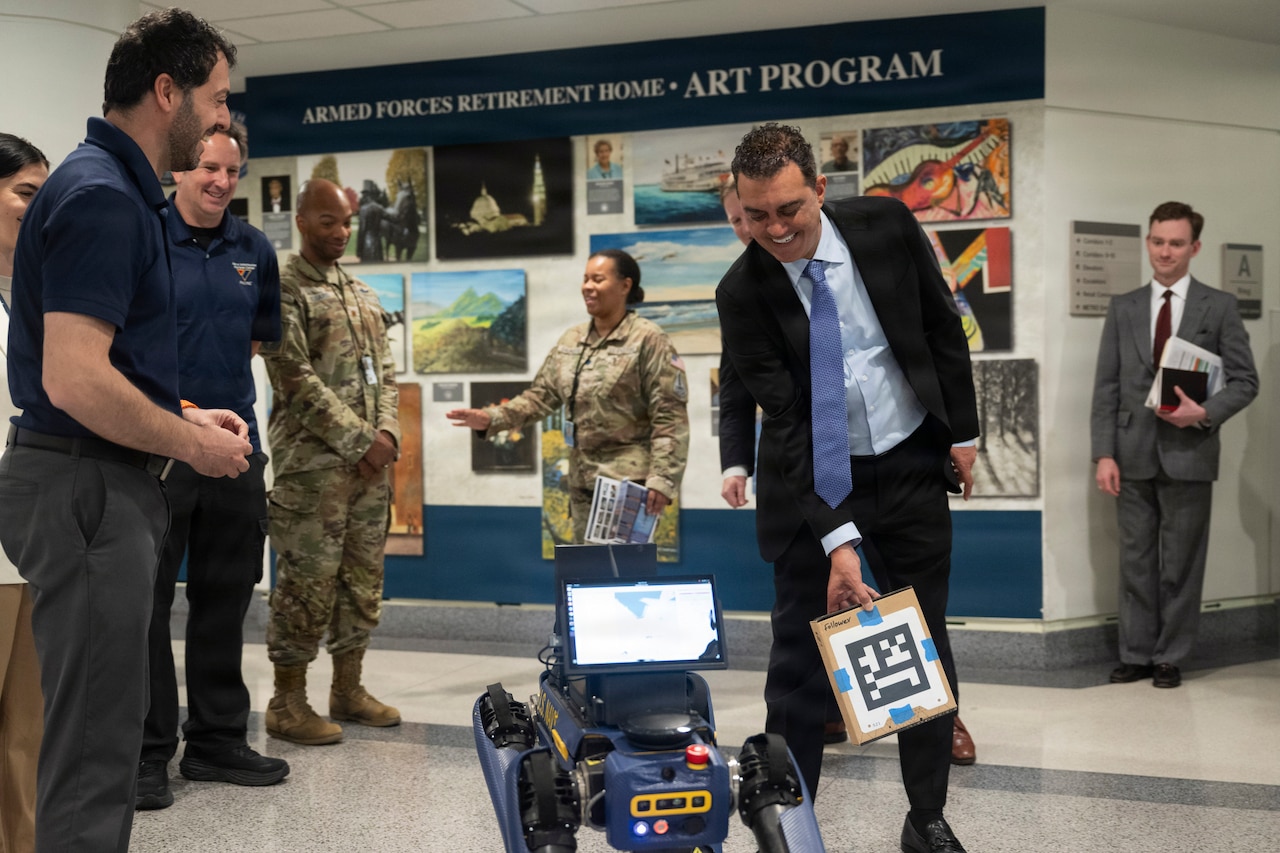 A person in a business suit holds a box with a QR code near a robotic dog inside a building, with other people looking on and posters on a wall in the background.