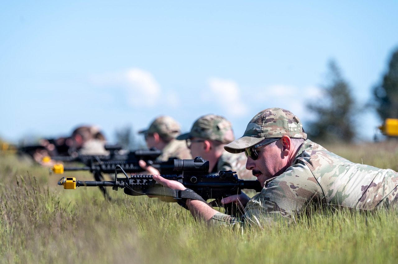 Four people in camouflage military uniforms lie in a row in tall grass under a blue sky with clouds and aim weapons.