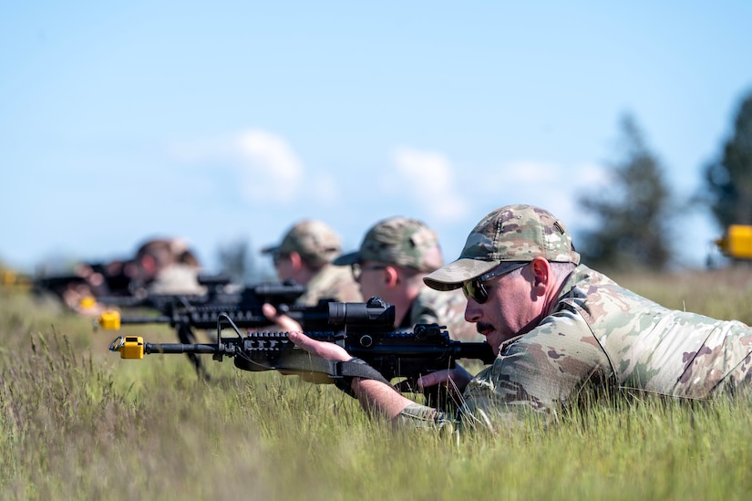 Four people in camouflage military uniforms lie in a row in tall grass under a blue sky with clouds and aim weapons.