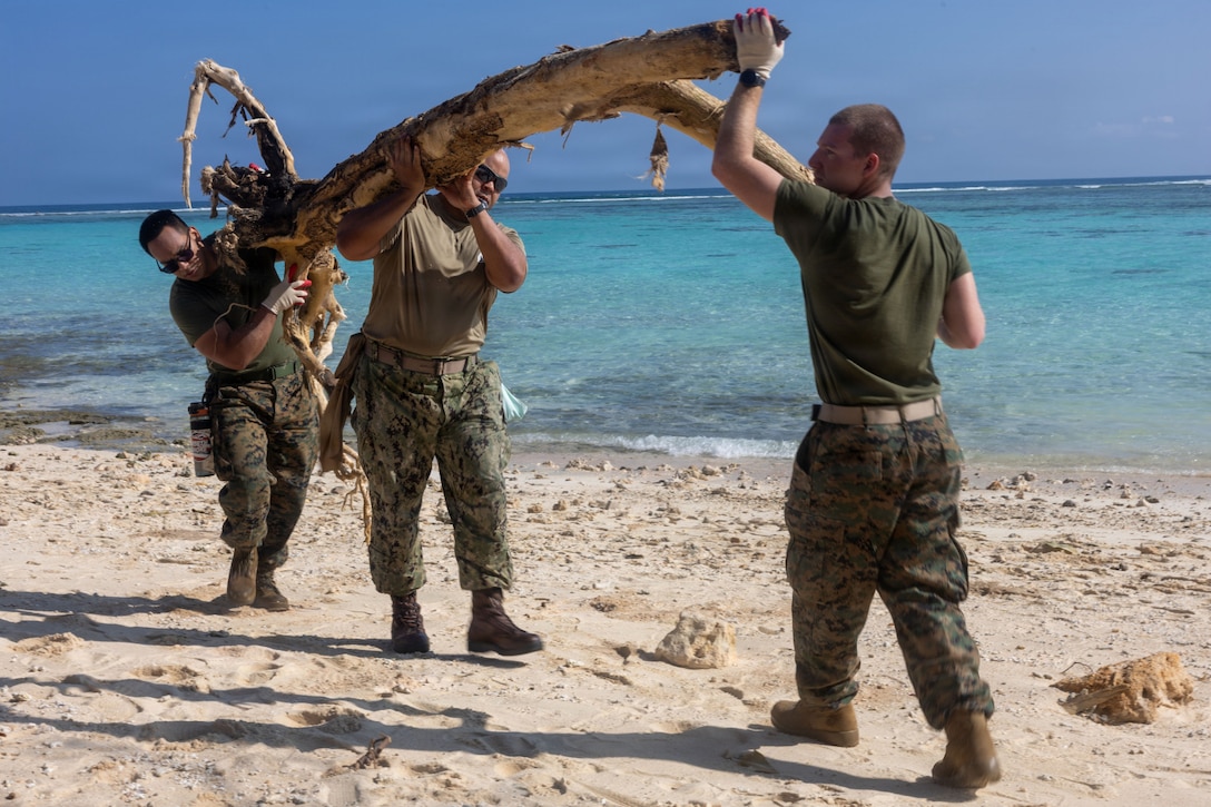 Three men in camouflage military uniforms move a large log on a beach.