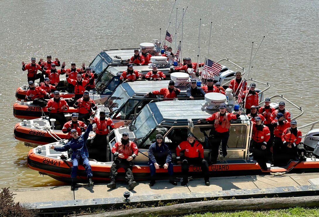 Four USCG Small-Boats with crew are tied up at a dock on a sunny day.  The crews, wearing cold water survival gear and lifejackets, look jubilant with high morale.