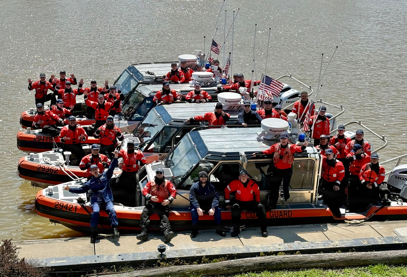 Four USCG Small-Boats with crew are tied up at a dock on a sunny day.  The crews, wearing cold water survival gear and lifejackets, look jubilant with high morale.