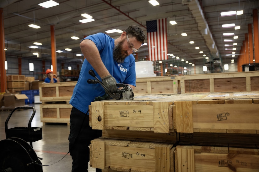 A man wearing casual attire uses a machine to attach bands to a wooden shipping box in a warehouse; there is an American flag hanging from the ceiling in the background.