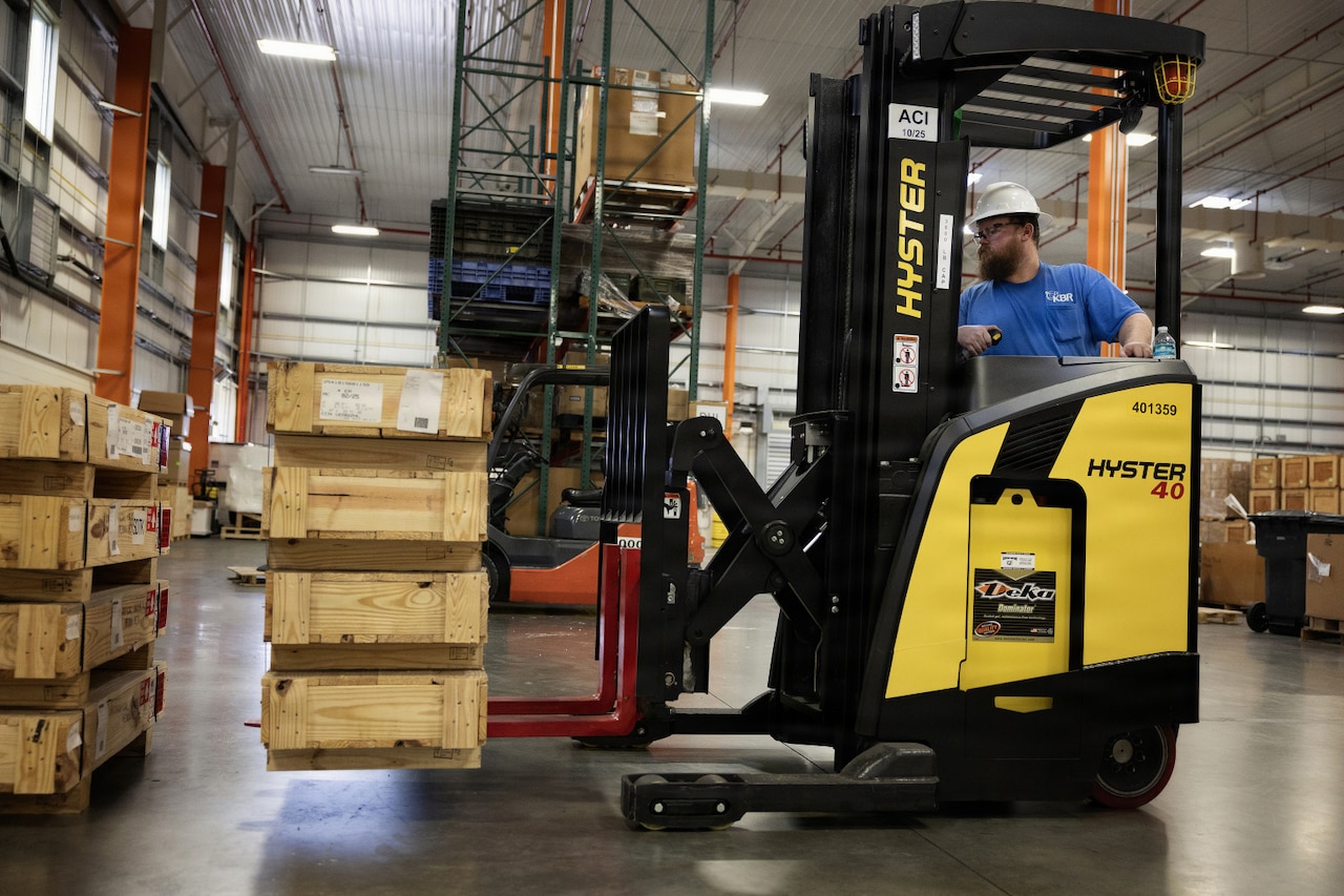 A man wearing casual attire uses a forklift to move wooden shipping boxes in a warehouse.