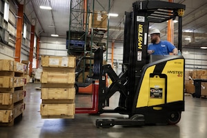 A man wearing casual attire uses a forklift to move wooden shipping boxes in a warehouse.
