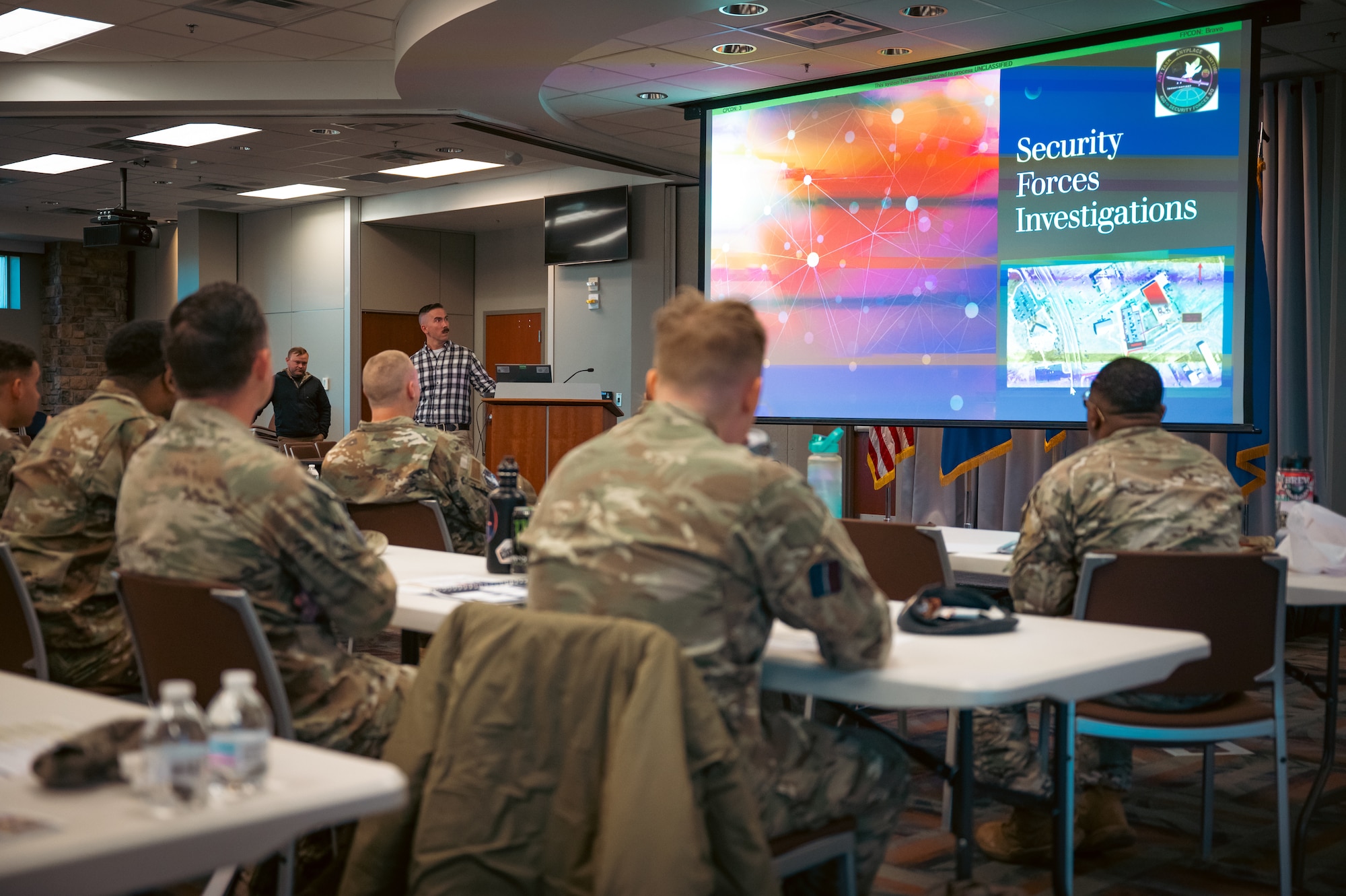 people sitting at tables in attendance with a projector screen showing briefing slides