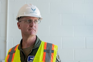 A man with glasses, wearing a white hardhat and a yellow construction vest, is positioned on the left side of the frame, looking to the right.