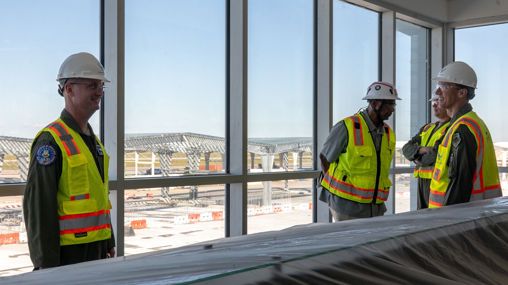 Four people wearing yellow construction vests talk in a construction zone. They appear to be on the second floor of a structure, standing in front of a large window. Out the window you can see construction of an aircraft hangar