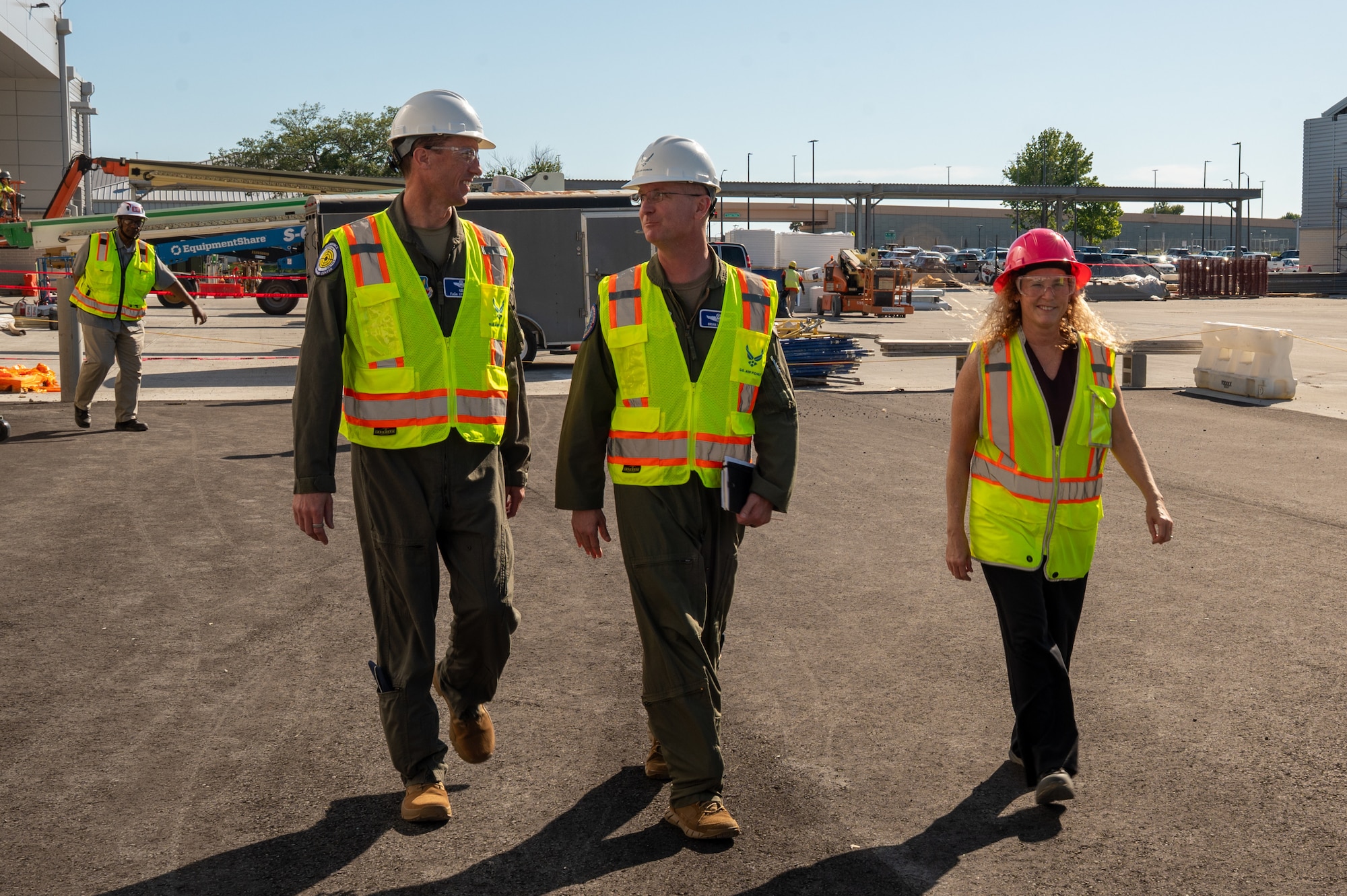 Three people in the foreground of the photo walk towards the camera, they appear to be talking amongst themselves. All are wearing construction vests and white hardhats. They are in a construction zone