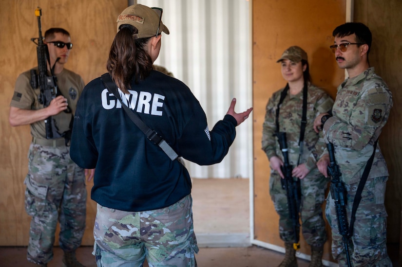 A woman in a camouflage military uniform and black jacket that says "CADRE" on the back speaks to three people in camouflage military uniforms who are holding weapons inside a training structure.