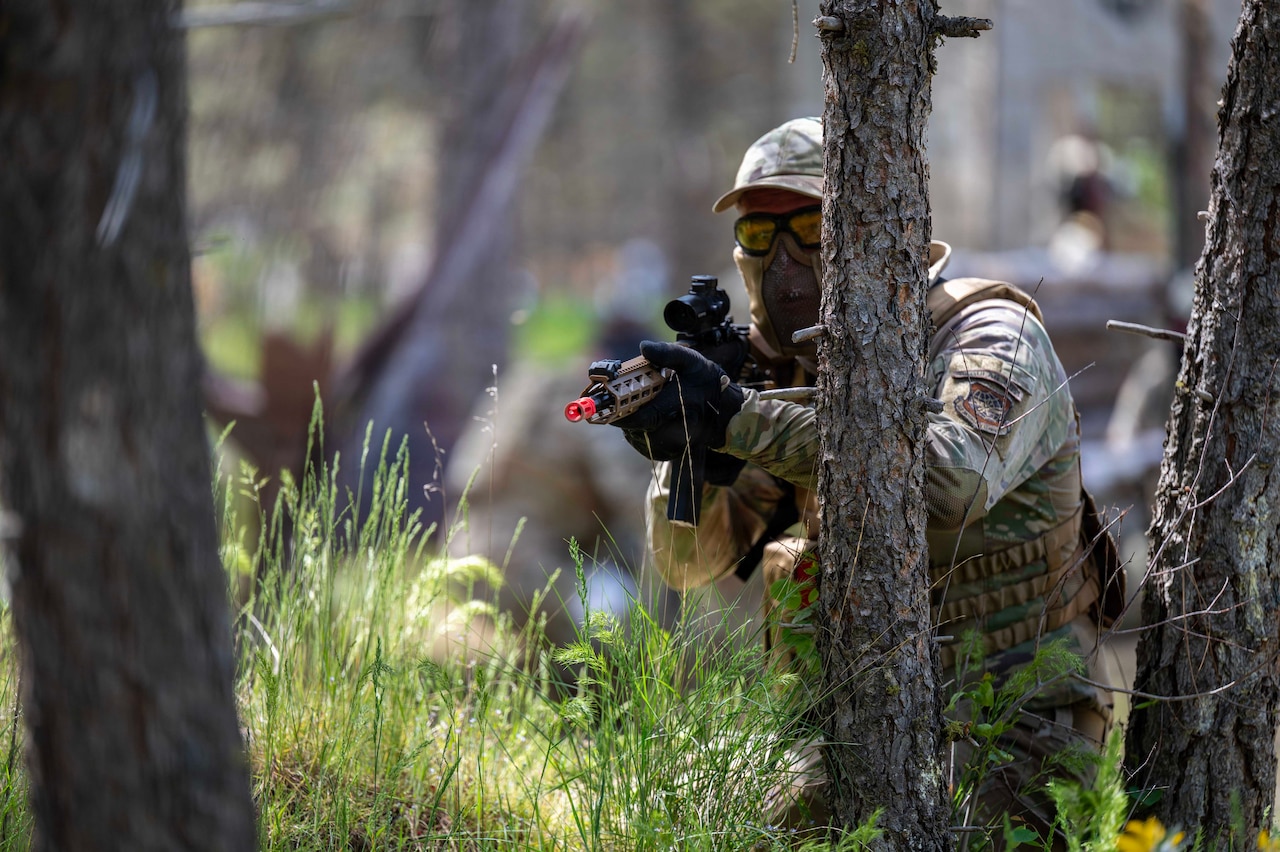 A person in a camouflage military uniform, goggles and a face covering stands in a wooded area and aims a weapon.