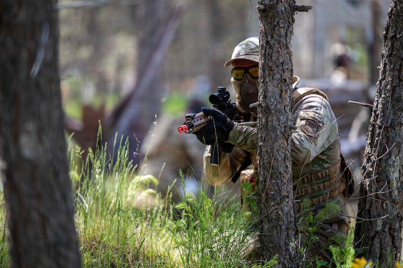 A person in a camouflage military uniform, goggles and a face covering stands in a wooded area and aims a weapon.