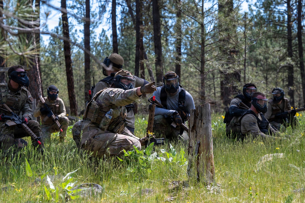 Eight people in camouflage military uniforms and face shields crouch in tall grass while holding weapons in a wooded area; one person points as the others observe.