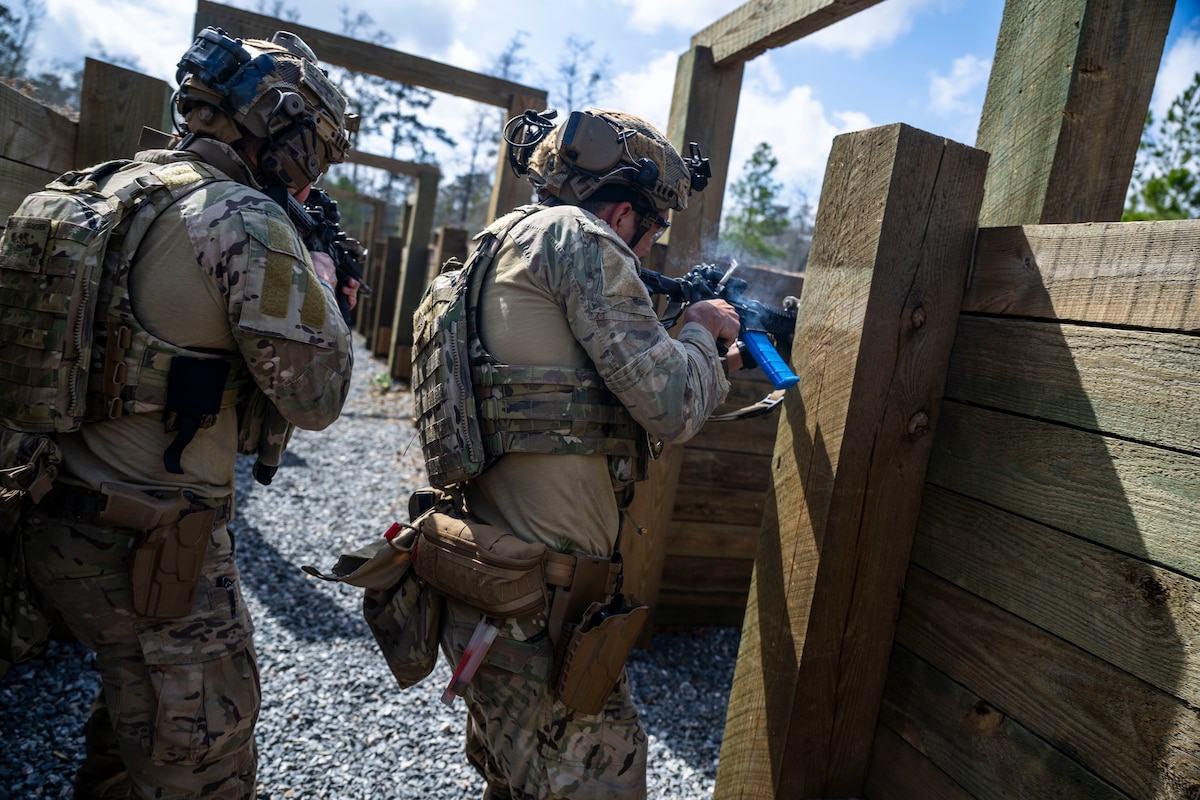 Two military members walk through a trench with weapons, one of them is firing his rifle around a corner
