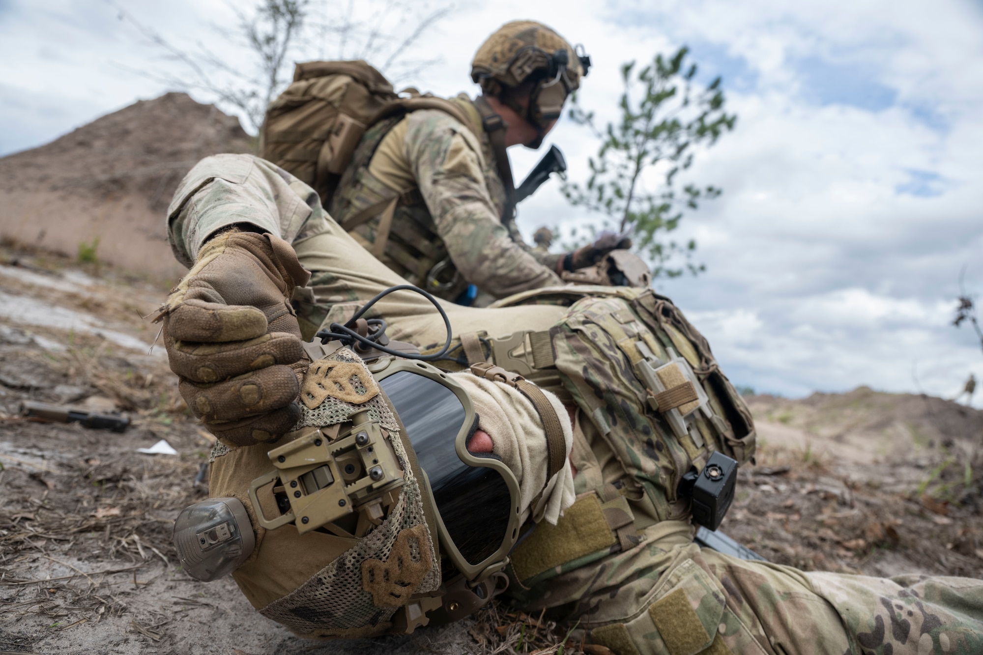 A military member wearing tactical gear lies on his back and looks at the camera, while another member simulates first aid