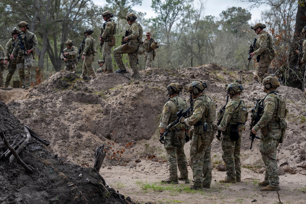 A large group of military members stand with rifles. Four of them are in the foreground and appear to be preparing to move. The others stand on a dirt mound behind them watching
