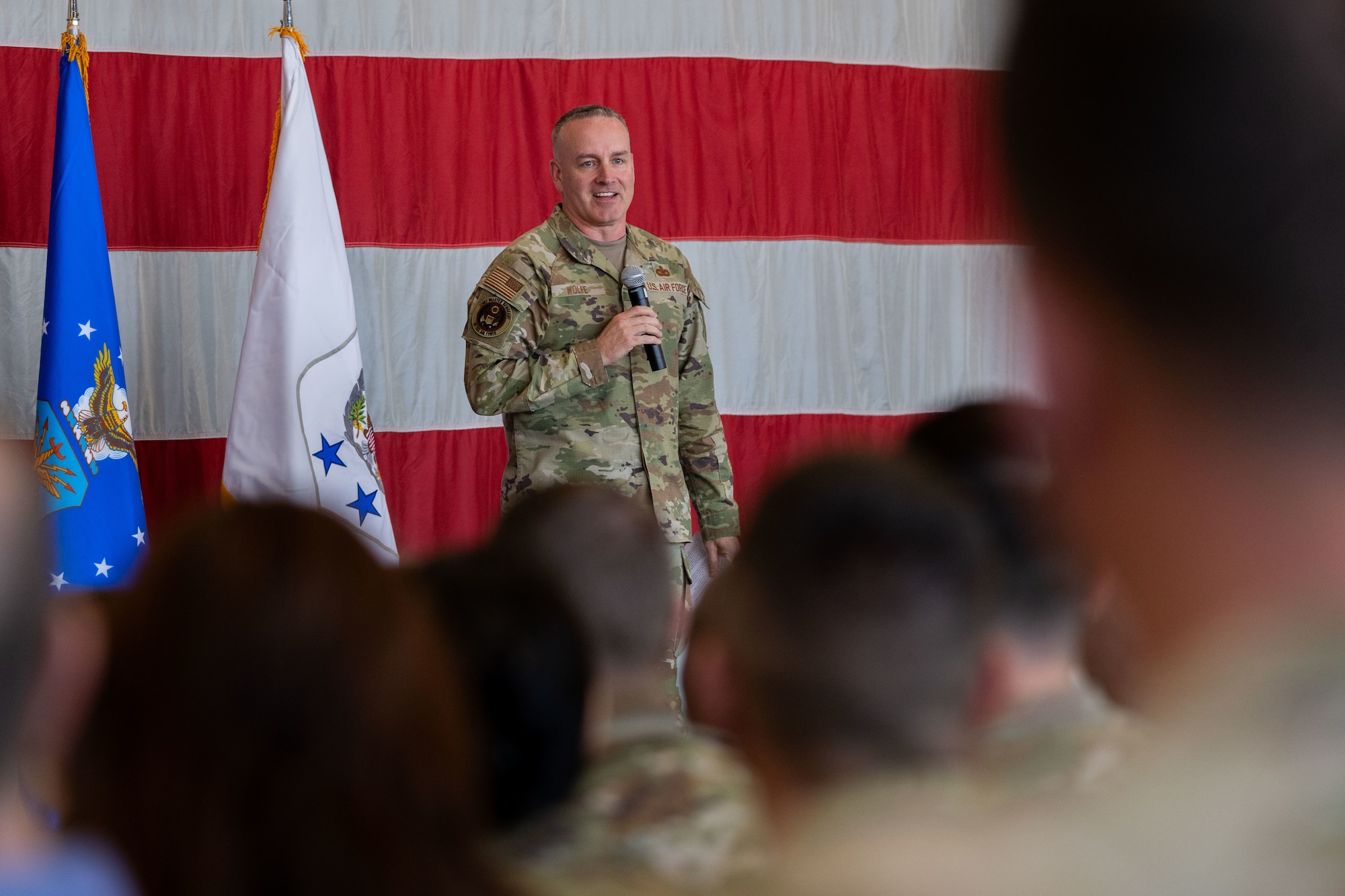 Air Force Chief Master Sergeant of the Air Force David Wolfe holds a microphone while addressing Airmen in the foreground of the image.