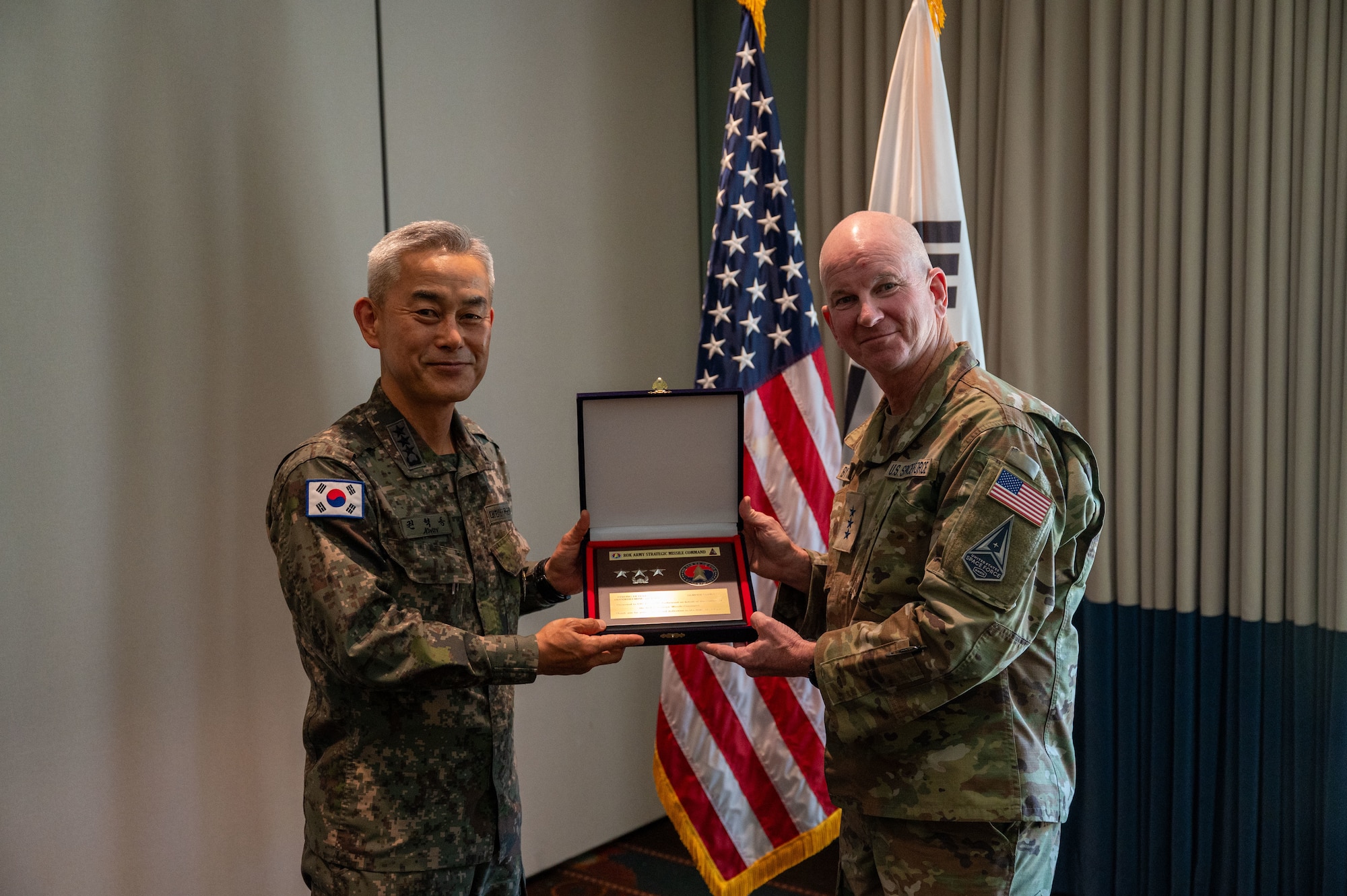An American and Korena man hold up a wooden box with a plaque inside and an embedded challenge coin