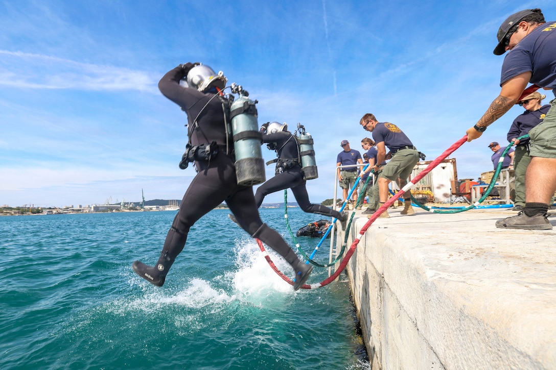 Two sailors in diving gear jump into blue water as personnel standing on a port hold a line connected to them during the day.