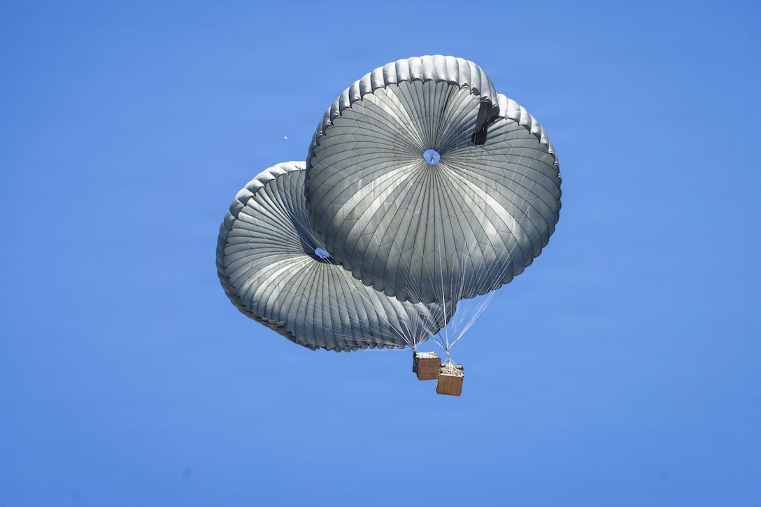 Two boxes descend with parachutes in a blue sky.