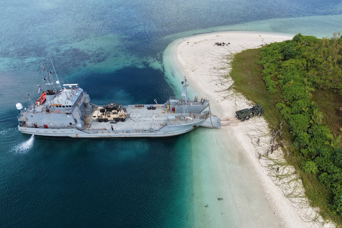 A military vehicle drives off a ship in blue waters onto an island during the day.